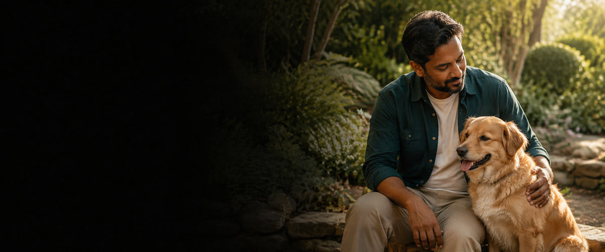 A man in a dark green shirt and beige pants sits outdoors with a golden retriever dog, set against a lush green garden background with a dark vignette on the left side.