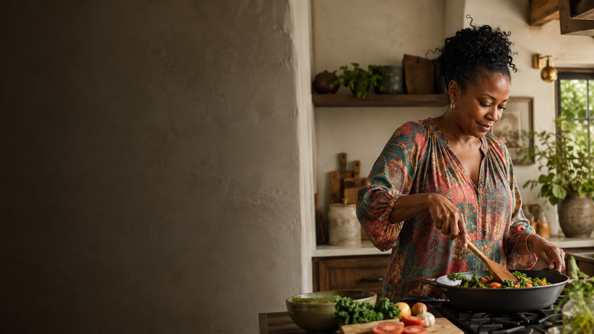 A woman with curly black hair wearing a colorful patterned blouse stirs a pan of vegetables in a rustic kitchen setting.